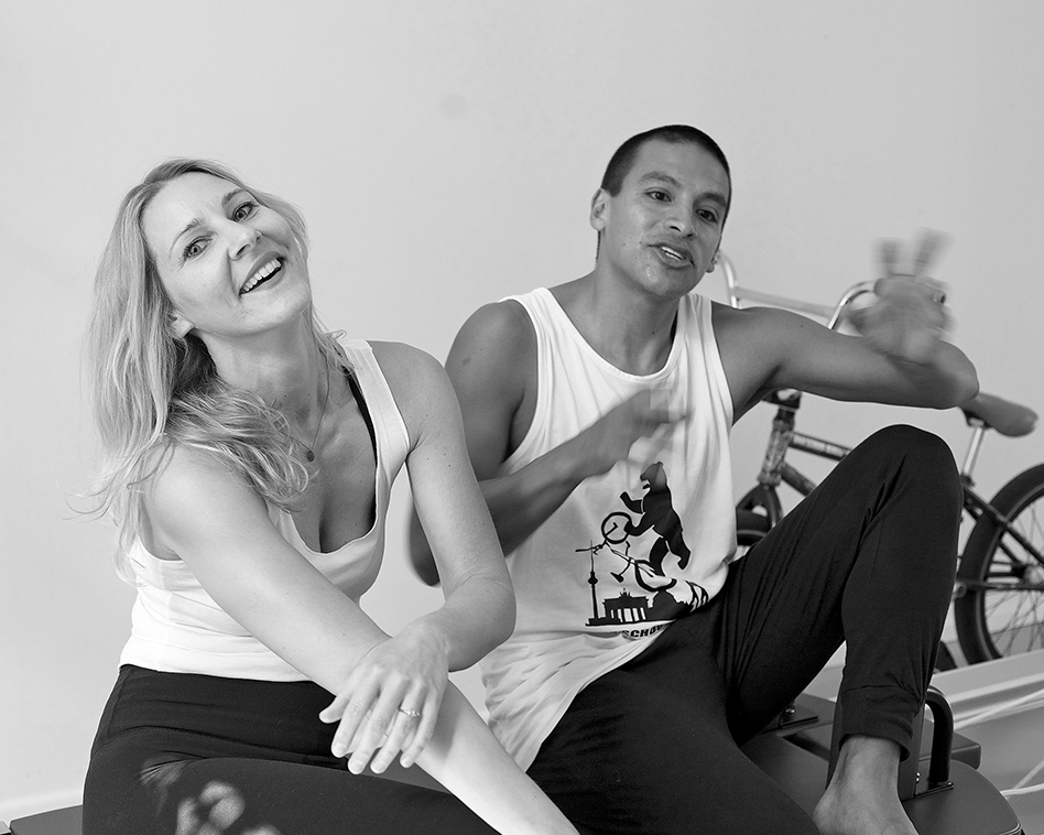 a portrait of two people, a woman laughs into the camera, her hair is tossed slightly back, her head is tilted and she crosses her arms, a man sits next to her and tells, while he moves his hands and one leg is upright in the background is a BMX bike