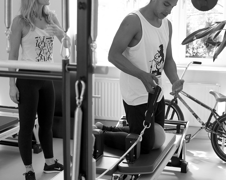 Solo Training im Pilates Studio, ein Mann bereitet sich auf dem Pilates Reformer für die nächste Übung, vor seine Trainerin steht hinter ihm