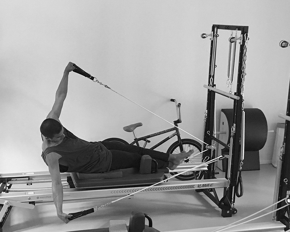 Pilates Übung, ein Mann trainiert auf dem Pilates Reformer, er hält mit beiden Händen die Handschlaufen und öffnet mit Federwiderstand seine Arme, der linke Arm zieht nach oben, der rechte Arm nach unten, sein Becken ist leicht nach hinten gekippt, seine Beine sind lang gestreckt, er hat viel Kraft