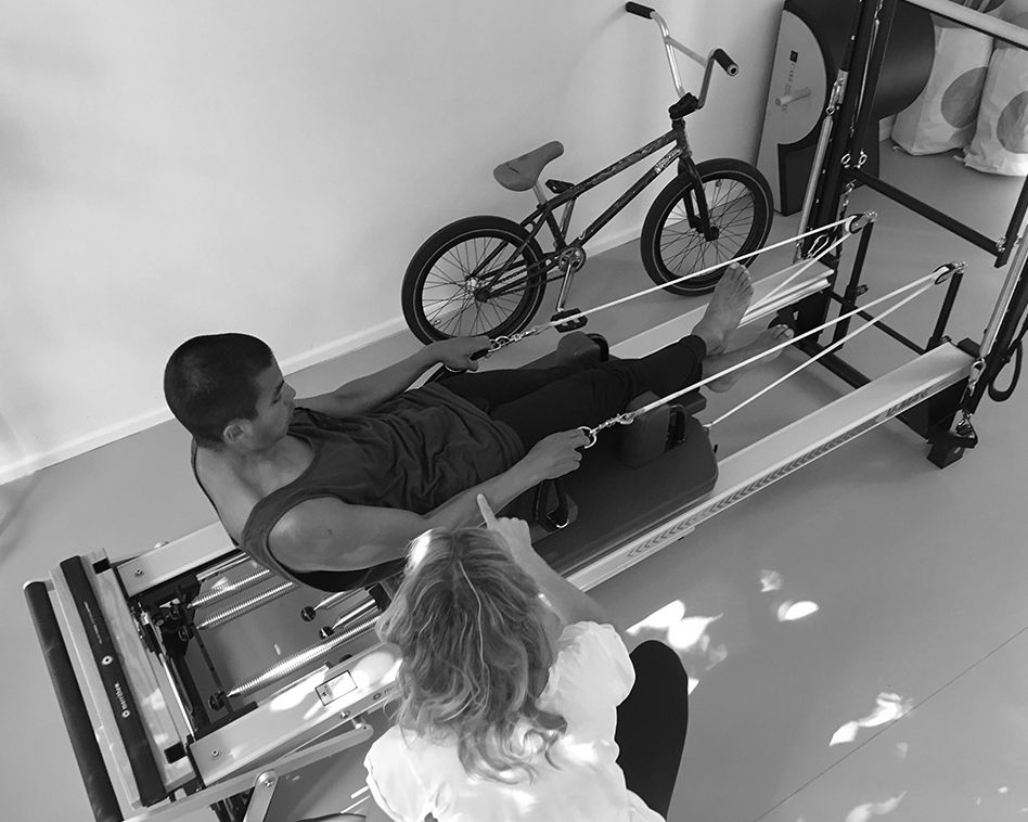 Pilates Training, ein BMX Sportler Trainiert auf dem Pilates Reformer, er sitzt und hält mit beiden Händen die Handschlaufen fest und rollt langsam mit seinem Becken nach unten ab, seine Pilates Trainerin sitzt neben ihm und leitet ihn genau an