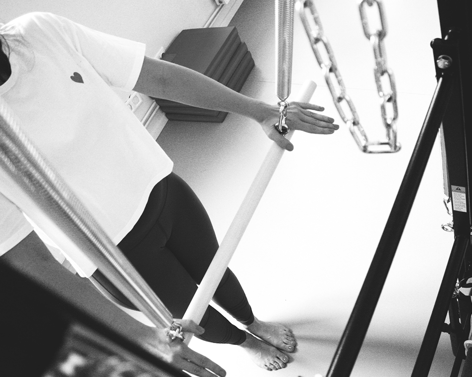 Pilates Studio, a woman is standing at the Tower Unit Wall, with a straight body alignment, she pushes the Tower Bare down with her hands to straighten her body and feel the length. The feet are closed