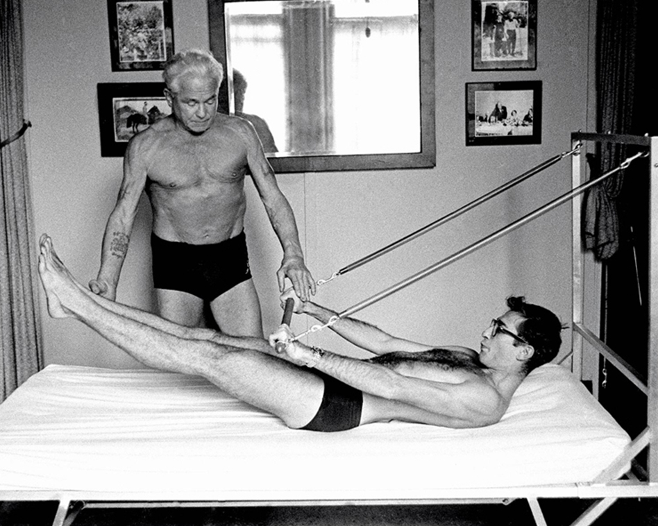 Pilates exercise, Jospeh Pilates training with a man Pilates. He lies on a bed and holds the Tower Barre firmly under spring tension, his upper body is slightly rolled up, his legs are 45 degrees long, his entire body is centered, Jospeh stands next to him and guides him.