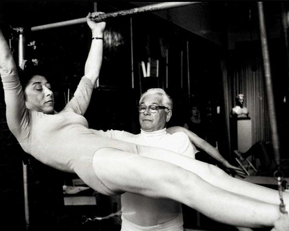 Jospeh Pilates with his client during training, he stands next to her and guides her, the client hangs with her body on the Pilates Cadillac, both arms grip the bar above, her feet are in the ankle strap, her entire body is powerful and centered.