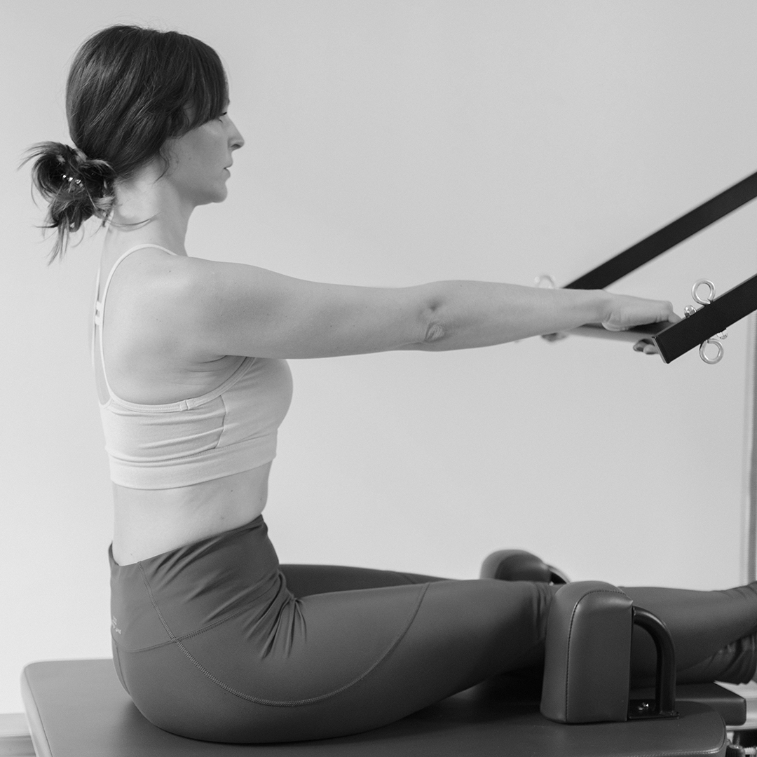 Pilates exercise, a woman sits straight up on the Pilates machine and holds the push-through bar with both arms, her back is straight and her posture straightened
