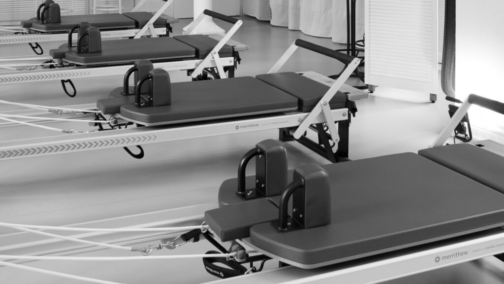 a Pilates studio with four Merrithew Pilates reformer machines standing next to each other, the frame is silver, the leather dark, the foot bar is pointed slightly upwards