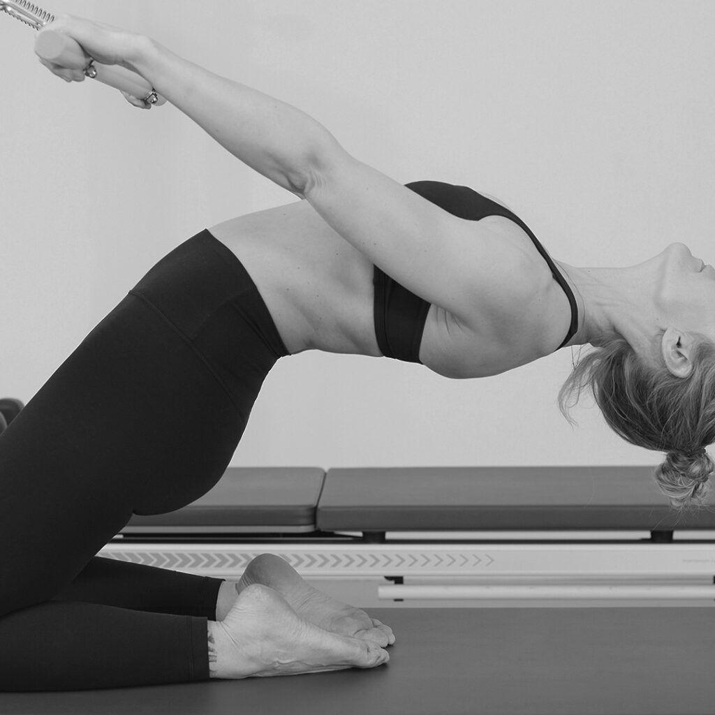 Pilates Übung, eine elegante Frau in sportlich, schwarzer Kleidung, lehnt sich genußvoll nach hinten und öffnet Ihren Oberkörper mit Hilfe der Zugkraft der Federn von der Pilates Tower Unit Bare.