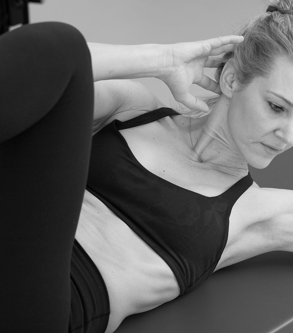 Close-up of a woman doing Pilates training, she has bent her knee to her body, her hands behind her head and is pulling her elbow to the knee, while she rolls up her upper body and tenses her abdominal muscles