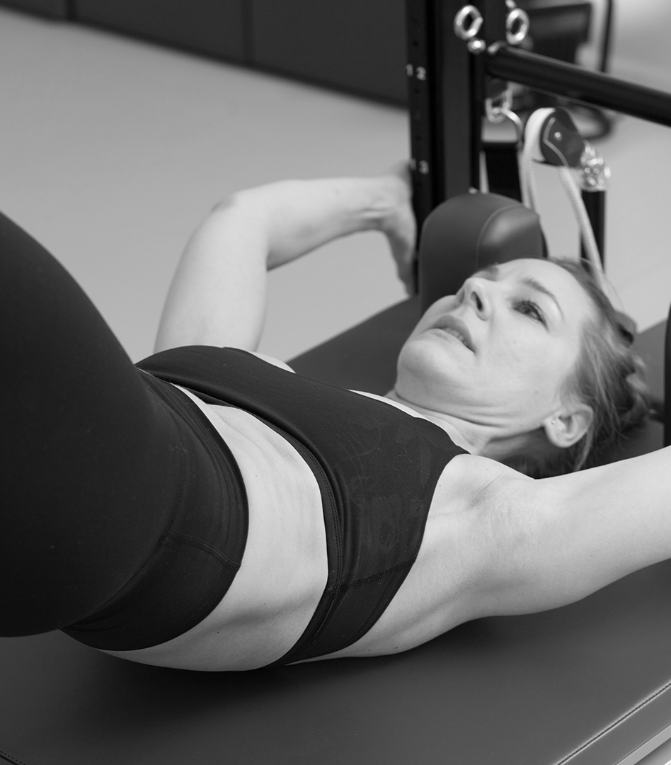 a young woman doing Pilates training at the Tower Unit Wall, her upper body is rolled up except for the shoulder blades, both hands hold the frame of the tower tightly, she holds herself up with strength