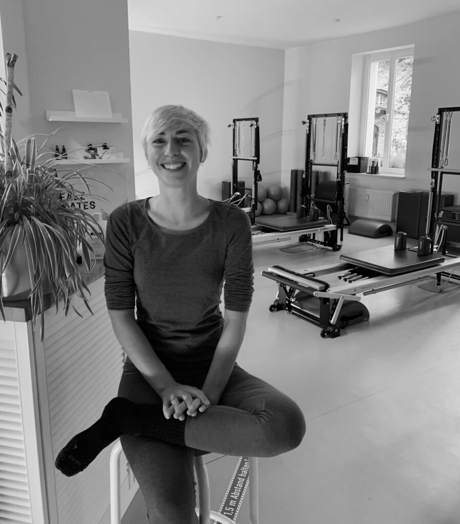 a young woman laughs straight into the camera, she is sitting in a Pilates studio, with a reformer and Tower Unit Pilates equipment in the background.She is happy.
