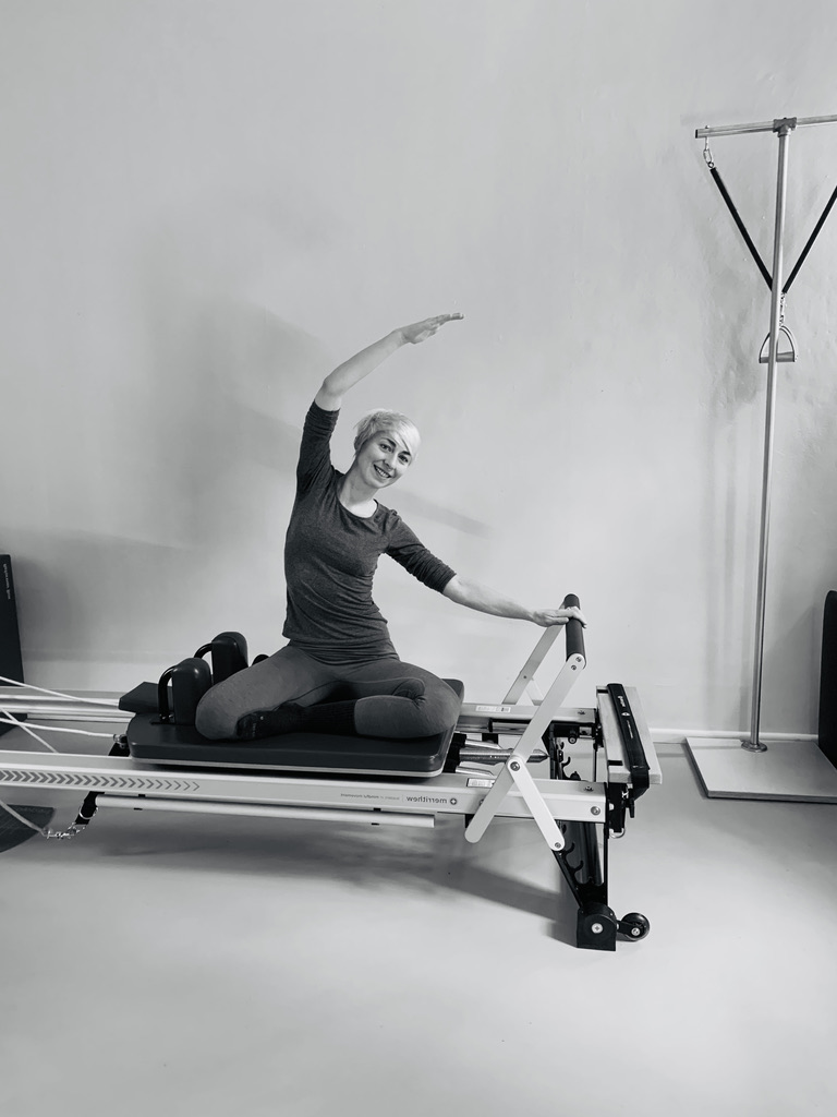 a young woman, trains on the Pilates reformer and shows the exercise mermaid, her arm pulls over her head, with the other arm she supports the foot bar away and she enjoys the lateral stretch.