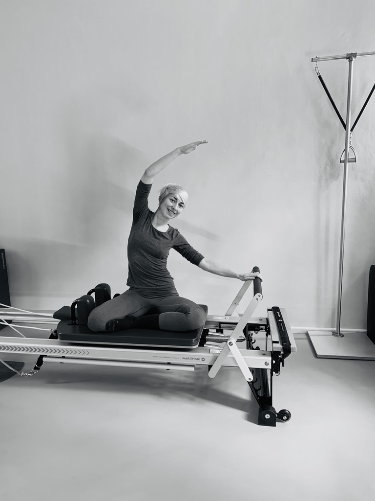 a young woman, trains on the Pilates Reformer and shows the exercise mermaid, her arm pulls over her head, with the other arm she supports the foot bar away and she enjoys the lateral stretch.