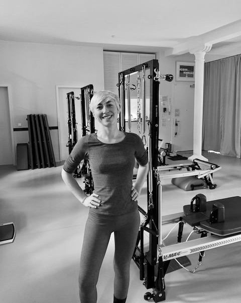 a happy woman standing confidently in the pilates studio. In the background there are Pilates reformer and tower unit equipment.