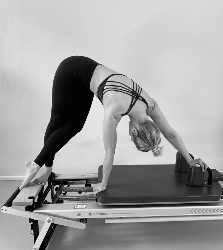An athletic woman on the Pilates reformer. Elegantly she goes into a side stretch. She wears black pants and body-hugging top. 10 reasons why now is the right time to start Pilates.