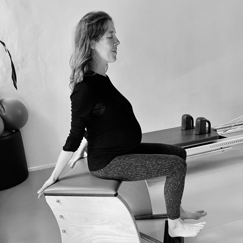 A pregnant woman sits on the Pilates chair and enjoys the stretching and straightening of her entire body. Pilates during pregnancy.