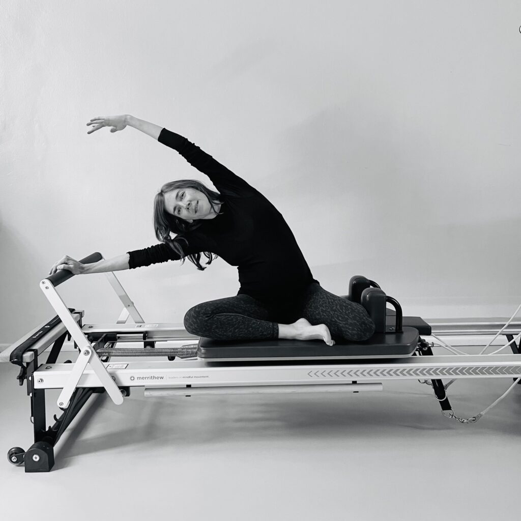 A pregnant woman stretches sideways on the Pilates Reformer and enjoys the side stretch. Pilates during pregnancy.