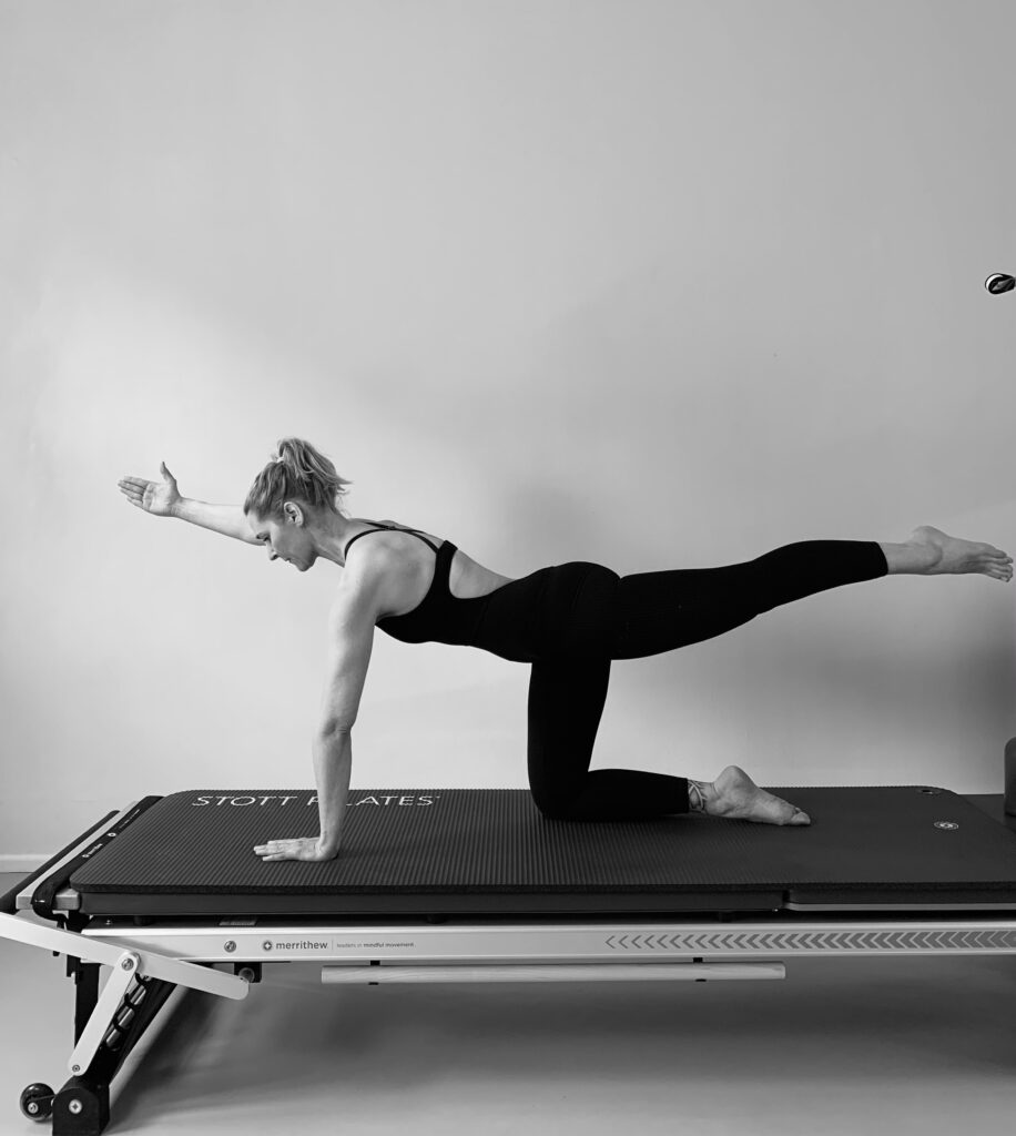a young woman stands on the Pilates Tower Mat and performs a Pilates exercise with one leg stretched back and one arm stretched forward