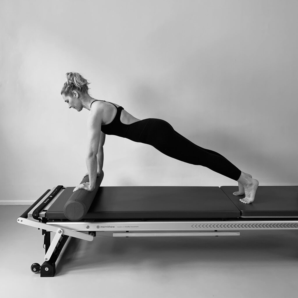 A young woman stands on the Pilates Tower Unit mat with the Foam Roller in a Pilates Plank exercise.