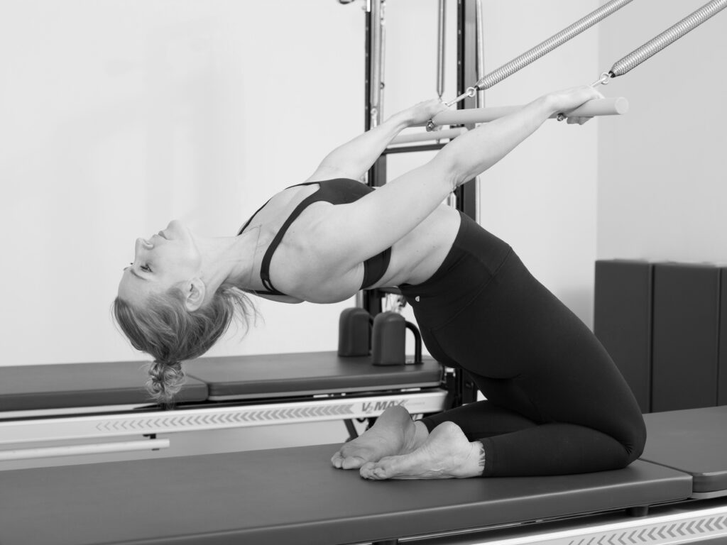 Eine junge Frau an der Tower Unit Wall, zeigt Pilates Übung zur Brustöffnung, Dehnung und Stabilisation der gesamten Körpermitte.