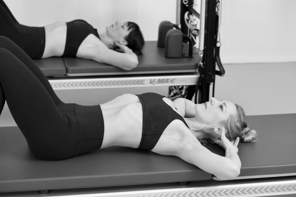 Two young women lying on the Pilates Tower Unit Wall in athletic clothing.