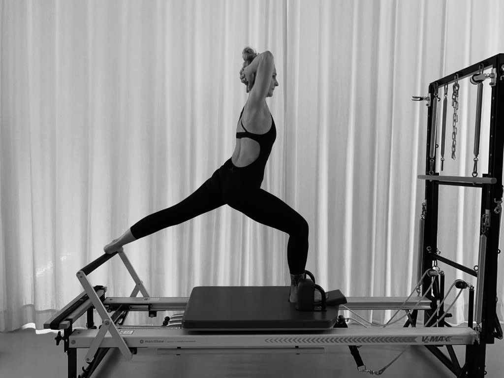 A young woman exercising on the Pilates Reformer in a standing position. She has her arms folded behind her head and is doing the Pilates exercise.
