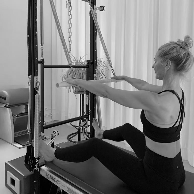 A young woman sits at the Pilates Tower Unit Wall with the barrel, upright, feet against the tower and she pulls the bar down. Body alignment.