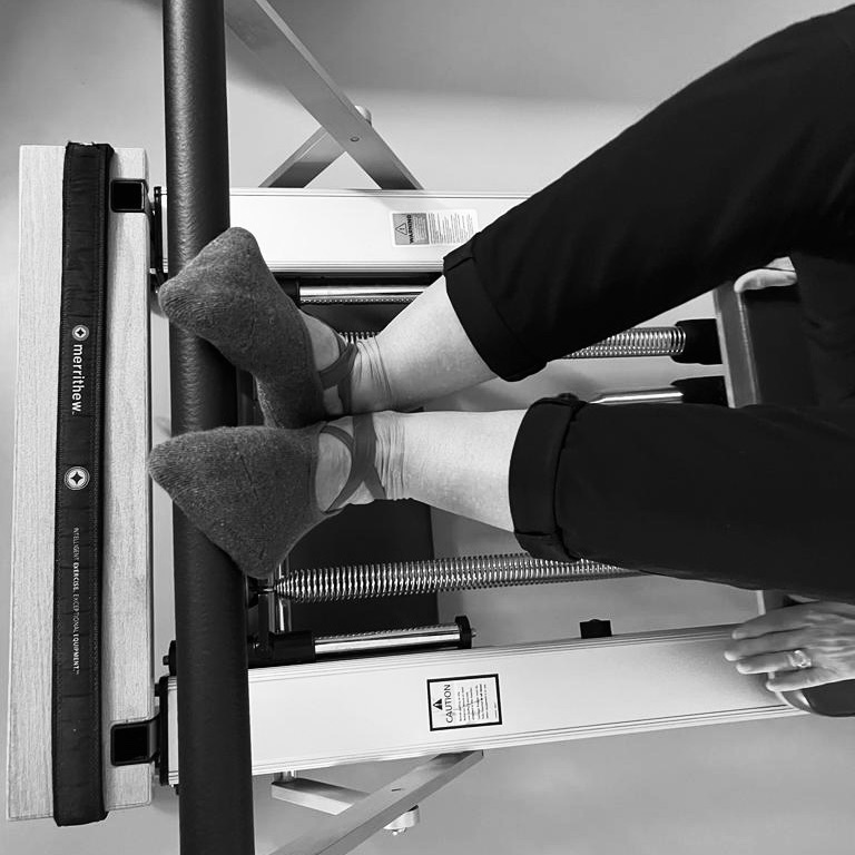 An older woman has both feet on the Pilates foot bar. In the Pilates V-position and lying with her back on the Pilates Reformer. Pilates in old age.