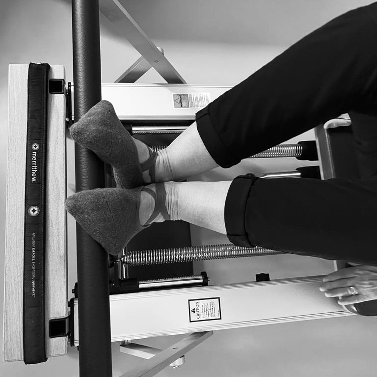 Eine ältere Frau hat beide Füße auf der Pilates Fußstange. In der Pilates V-Position und liegt mit dem Rücken auf dem Pilates Reformer. Pilates im Alter.