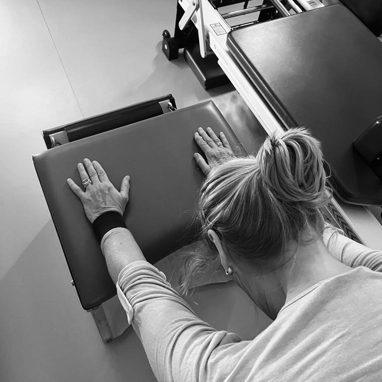 A woman holds both arms on the Pilates chair and goes into a stretching exercise. She is dressed athletically surrounded by Pilates equipment elements. Pilates in old age for optimal health.