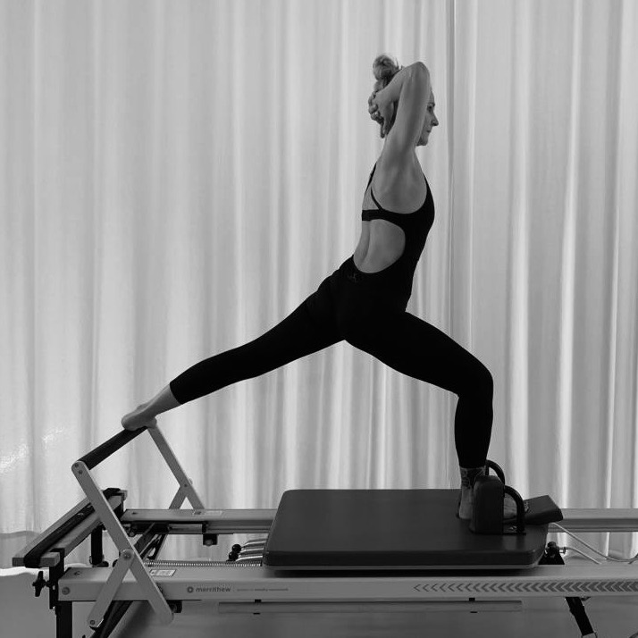 A young woman is exercising on the Pilates reformer. Her left leg is resting on the foot bar, her hands behind her head. Slowly she goes into a stretch. Pilates split.