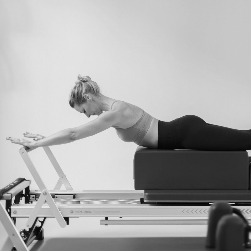 A young woman lies on the Pilates reformer, looking down. Her arms are stretched out and supported by the footbar. The entire body is lengthened. The physical stiffness is released.