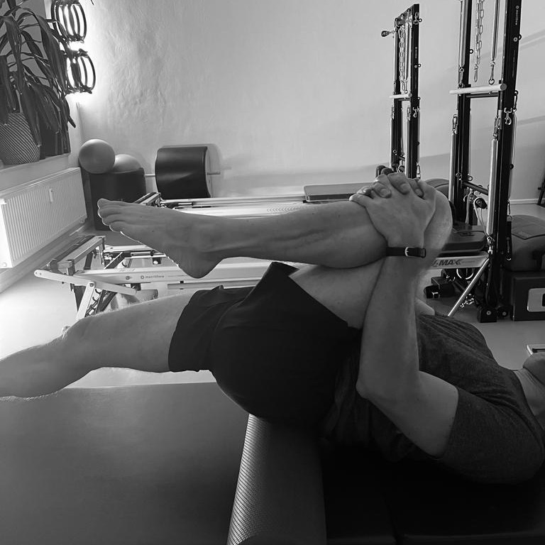Pilates Stretch für den Mann, umd ie Flexibilität zu stärken und Dehnung tief zu spüren. Ein Knie zieht fest an den Oberkörper, das andere Bein ist gestreckt.