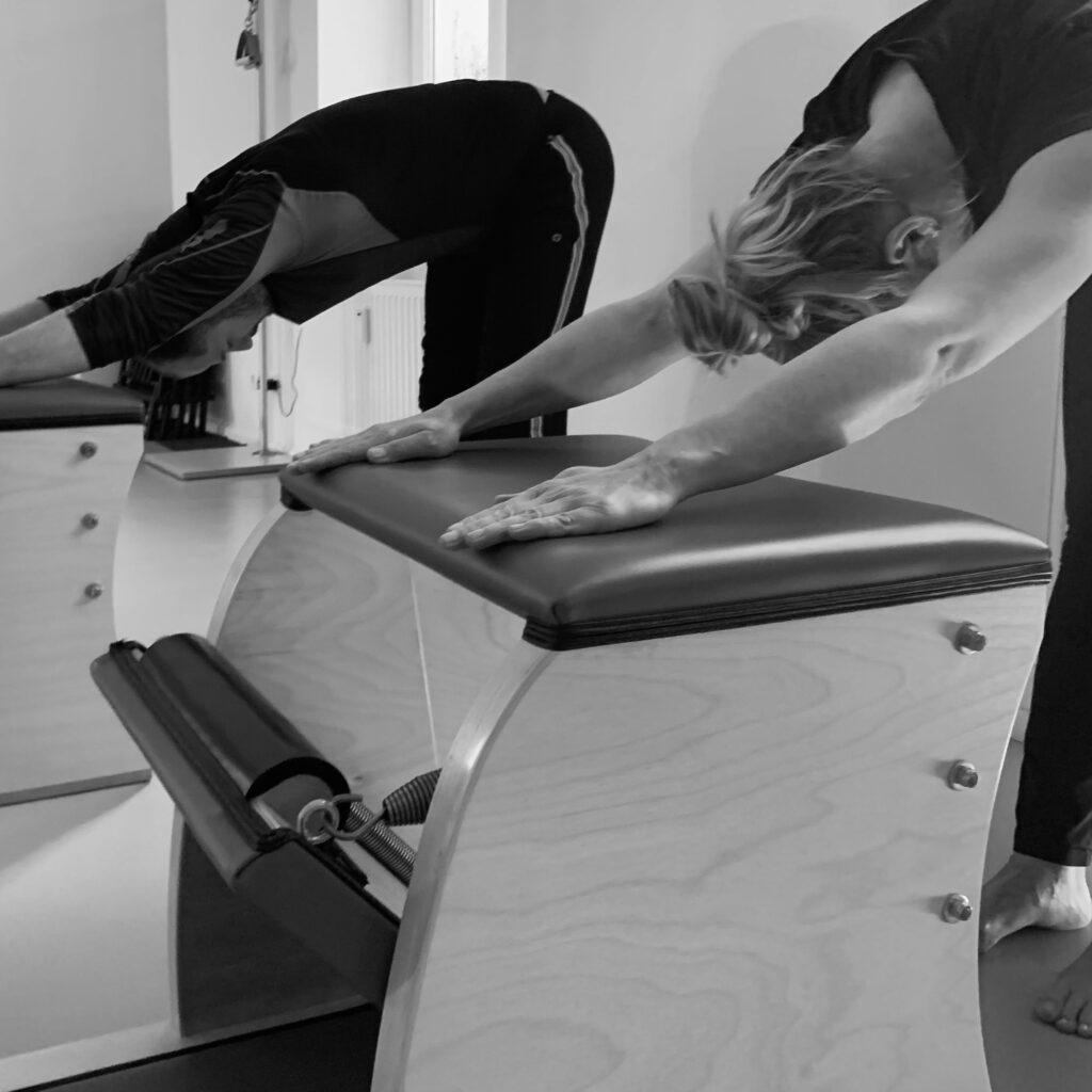 Two clients, a woman and a man, are training on the Pilates Chair and enjoying the wonderful stretch at the end of the class, their hands are placed far forward on the Pilates Chair, their heads are pointing downwards.