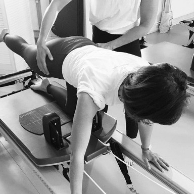 Pilates solo training on the Pilates Reformer. A young woman trains targeted hip exercises with the guidance of the Pilates teacher. The Pilates teacher corrects her and provides support.