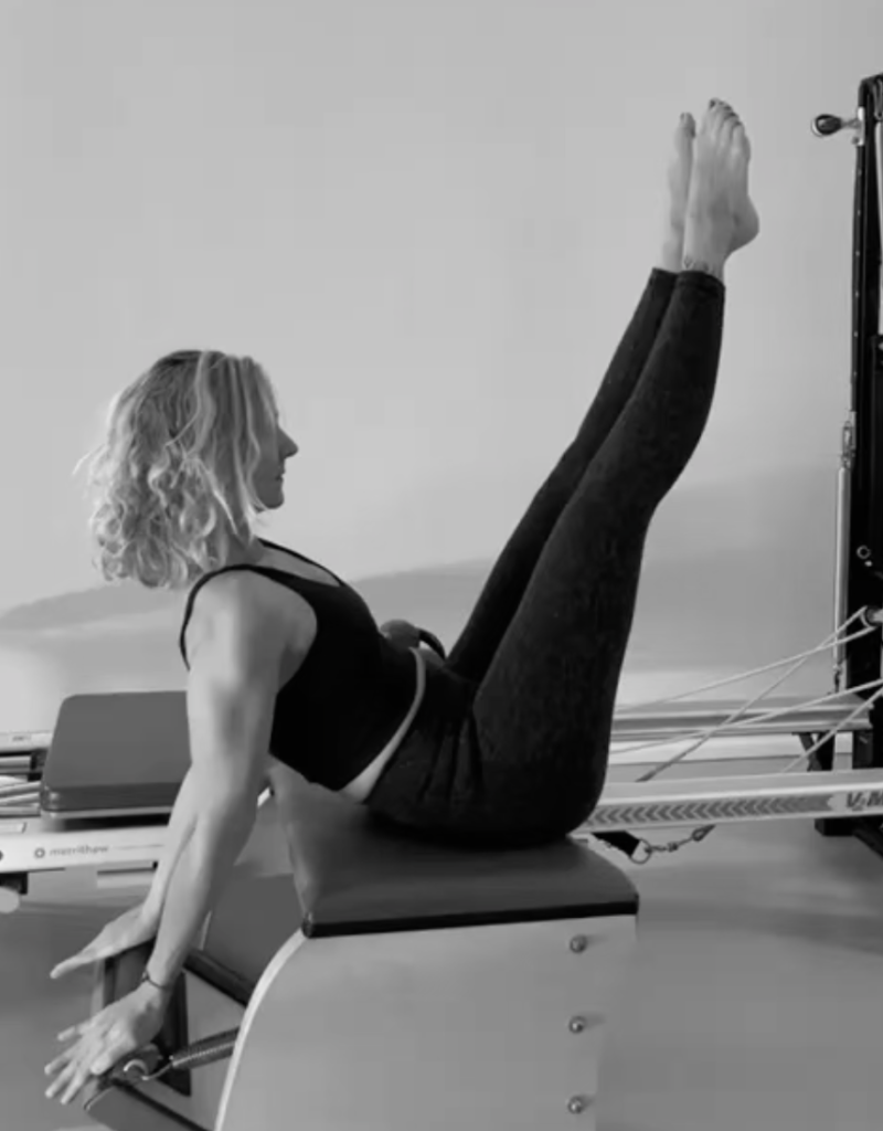 A young woman is exercising on the Pilates chair. Her arms are stretched out on the pedals, her legs are stretched upwards into the air. The entire balance comes from the centre of the body. She uses her strength to keep herself centred on the Pilates chair.