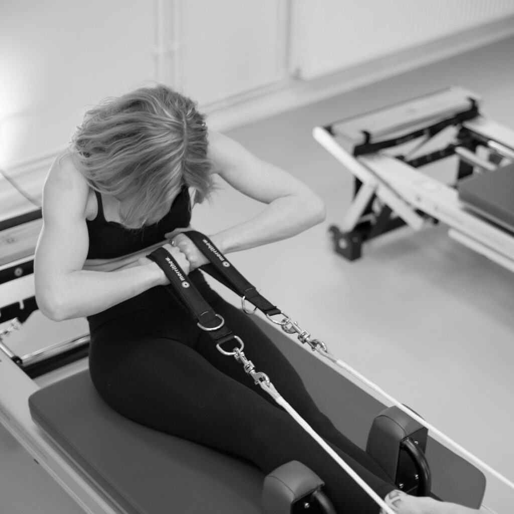 A young woman is training on the Pilates reformer, sitting on the sled with her legs crossed. Her arms are crossed in front of her chest with the loops in her hands. She rolls backwards, looking down towards her stomach.