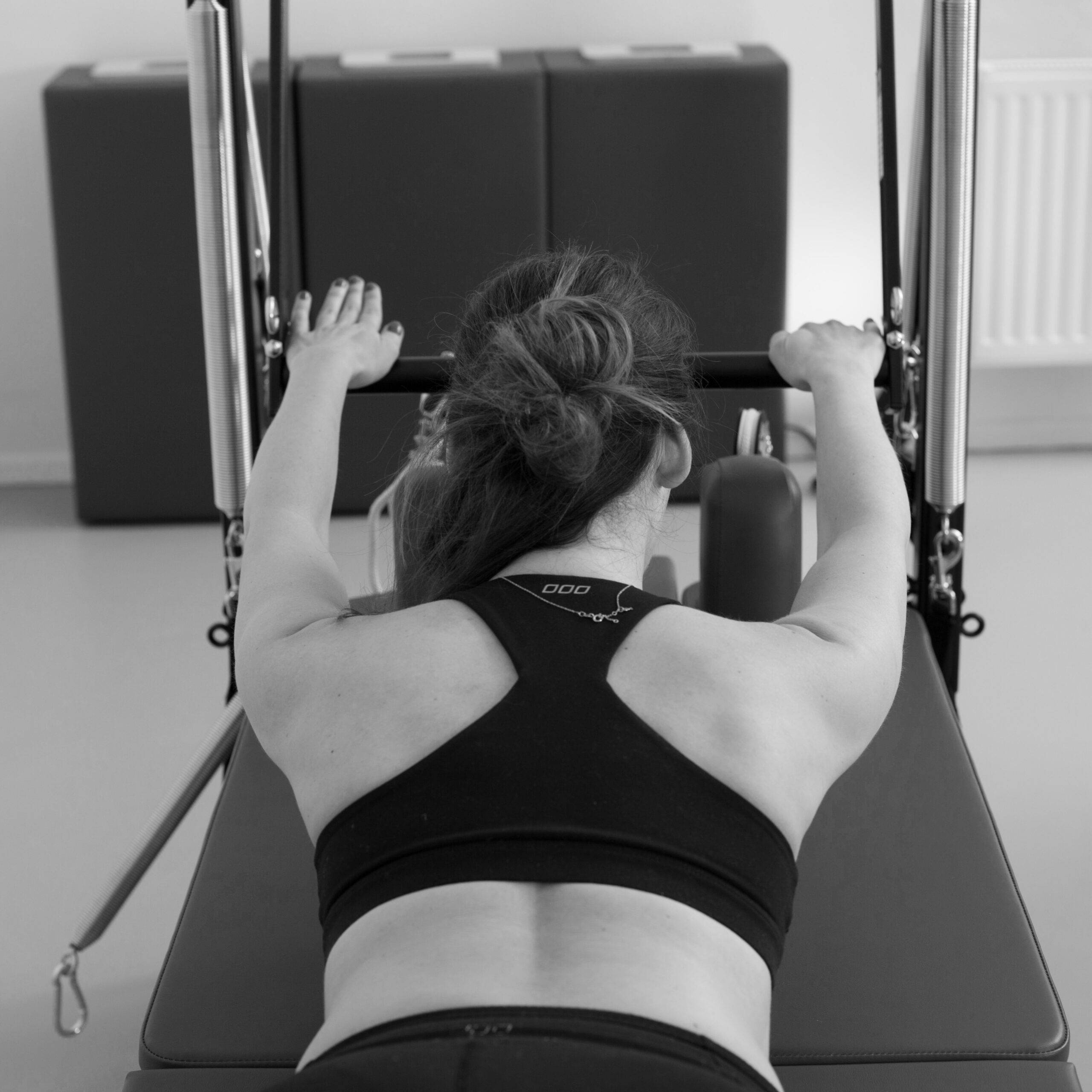 Eine junge Frau trainiert an der Pilates Tower Unit Wall. Sie liegt mit dem Bauch auf der Matte, ihre Arme sind gerade auf der Push-Throughbar abgelegt.