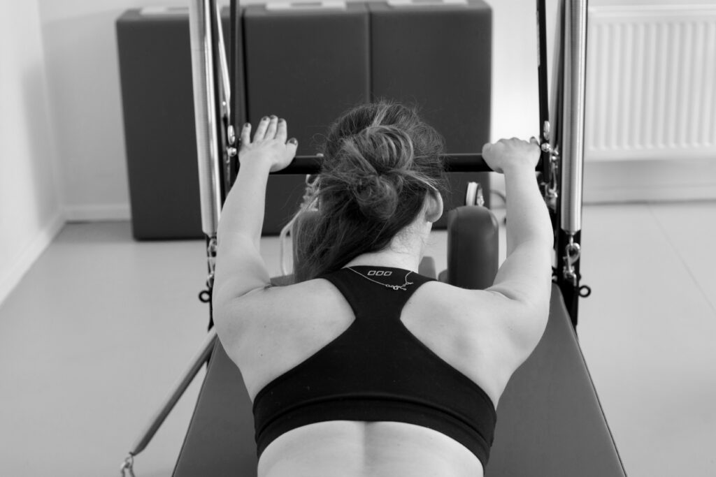 A young woman is training on the Pilates Tower Unit Wall. She is lying on her stomach on the mat, her arms are straight on the push-through bar.