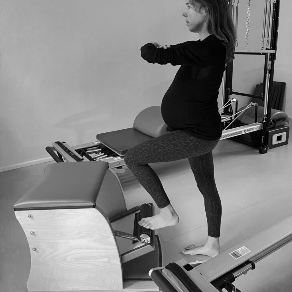 Eine schwangere Frau trainiert am Pilates Chair. Ihr einer Fuß ist auf dem Pedal, ihre Arme vor sich verschränkt. Die Übung wird im Stand ausgeführt.
