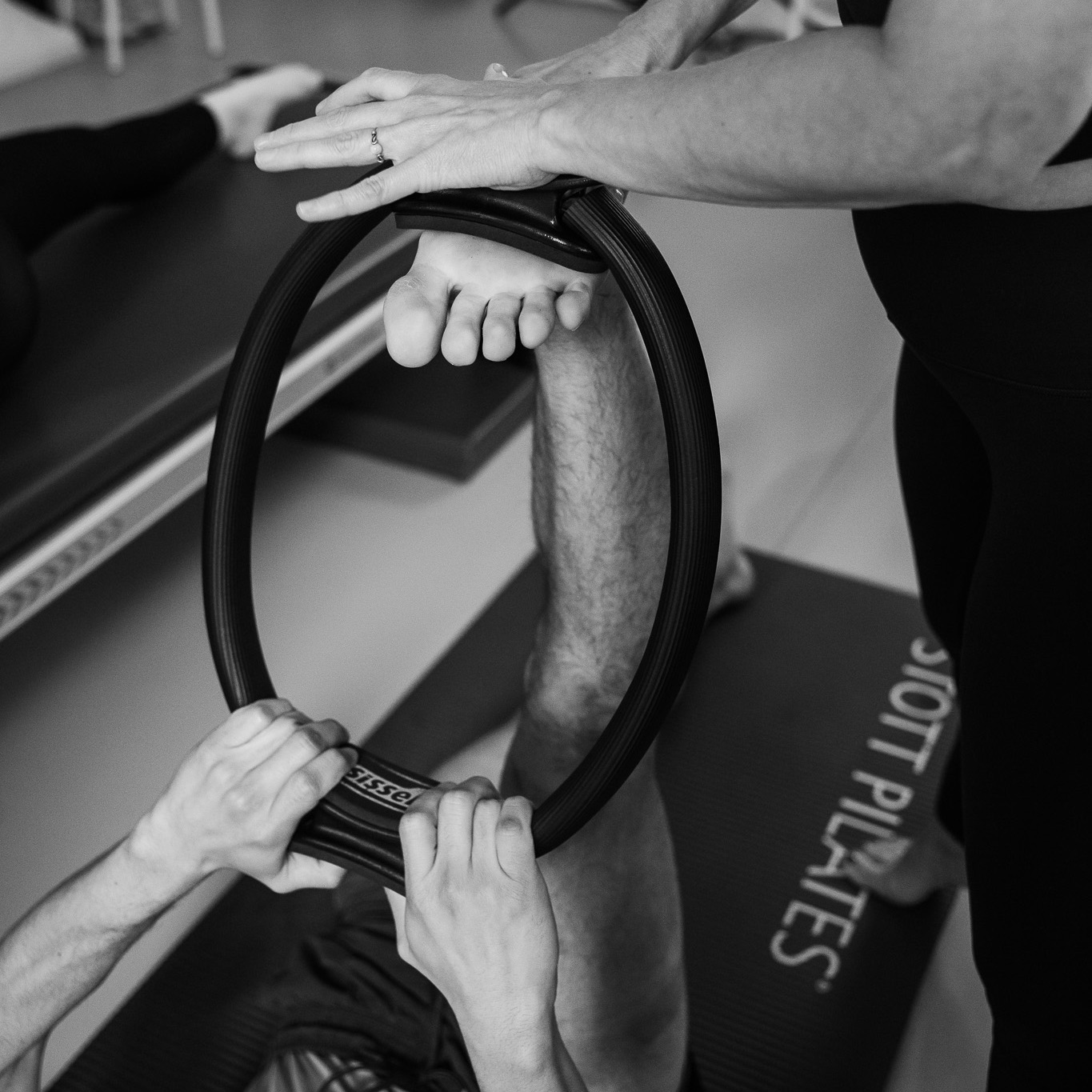 A young man lies on the Pilates mat with his foot in the Pilates Magic Circle and holds it with both hands. He stretches his entire leg and foot and enjoys the stretch. A Pilates teacher supports him during the intensive stretch. Get yourself and your body moving.