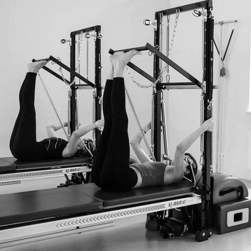 Two young women exercising on the Pilates Tower Unit. The legs are tucked in and the feet are on the push-through bar. Stretching the entire back of the legs to release stress and blockages.