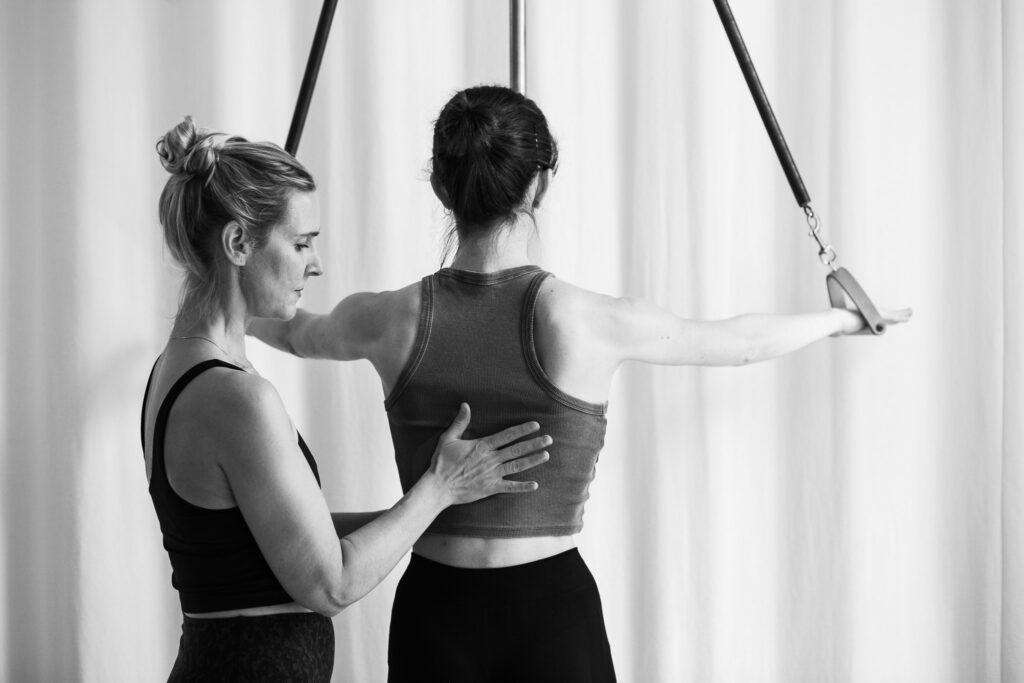 Strengthen the lower back. Become pain-free with regular Pilates training. A young woman strengthens her back muscles on the Pilates Pedi-Pole, both arms stretched out to the side with clear and tactful guidance from the Pilates teacher. She corrects the posture of her back. Pilates exercises to prevent a slipped disc in the lower back.