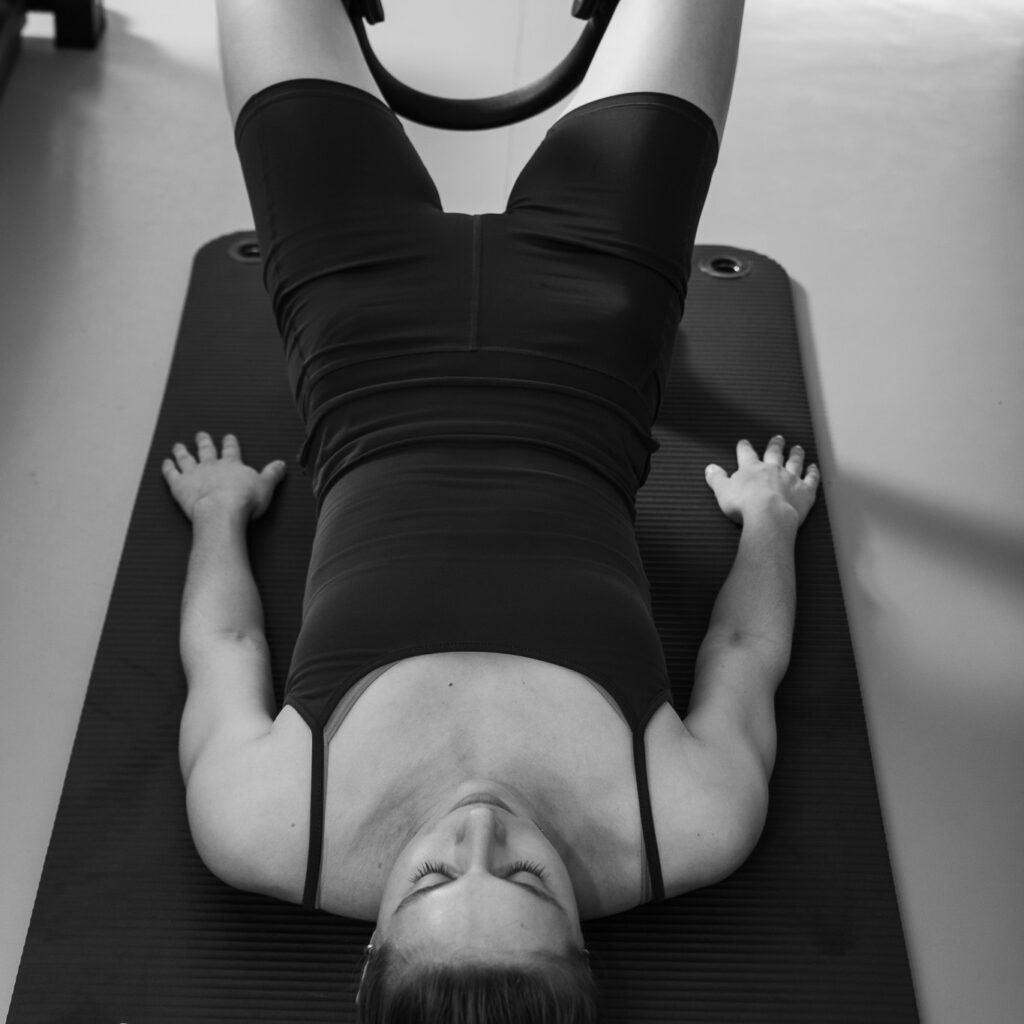 Beckenbodenmuskulatur stärken mit Pilates. Eine junge Frau liegt mit ihrem  gesamten Körper auf einer Matte, die Arme neben ihr und den Magic Circle zwischen ihre Beine. Eine Pilates Übung um die Beckenmuskulatur zu stärken.