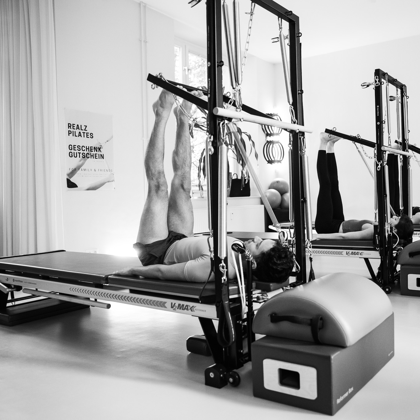 Footwork im Pilates. Ausgeführt an der Pilates Tower Unit Wall im Pilates Studio. Ein junger Mann liegt an der Tower Unit, seine Beine gestreckt, die Füße an der Push Through Bar gestreckt. In der Gruppenstunden werden Fußübungen durchgeführt.