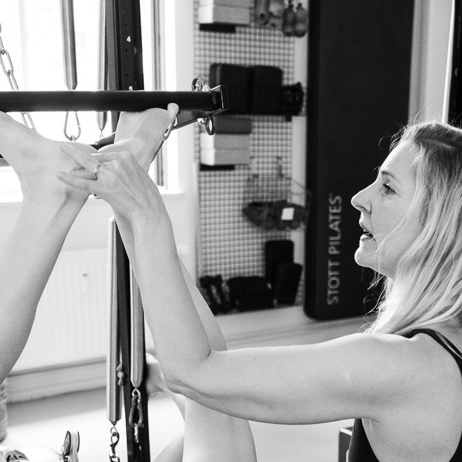 Pilates Footwork an der Pilates Tower Unit Wall. Die Zehen befinden sich an der Push-Through Bar und drücken den Federwiderstand weg. Eine Pilates Lehrerin sitzt daneben und korrigiert die Fußhaltung und präzise Ausführung der Fußarbeit im Training. Sie hält die Hacken zusammen. 