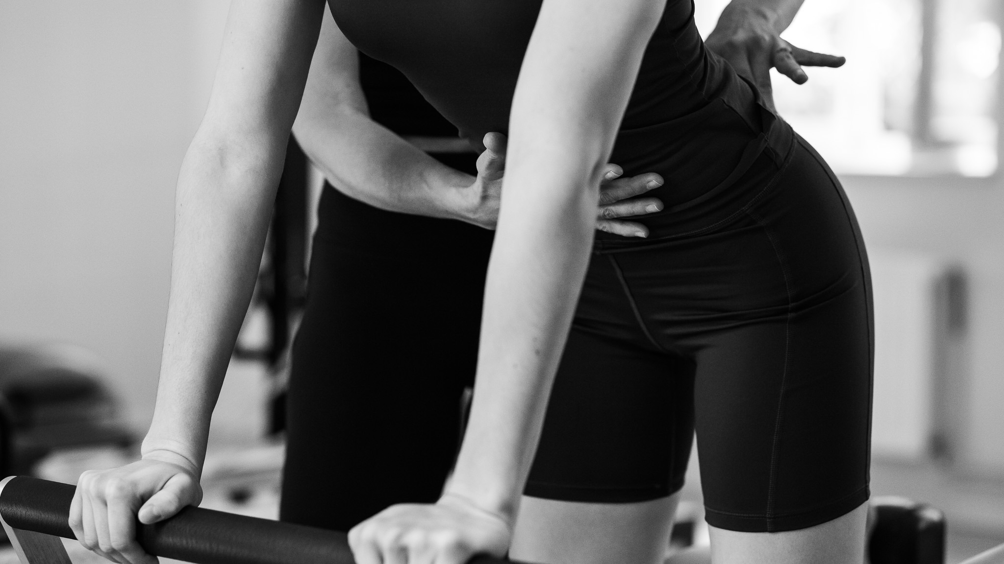 A young woman trains on the Pilates reformer. A Pilates instructor guides her and pays attention to her pelvic position to train the deep pelvic floor muscles.