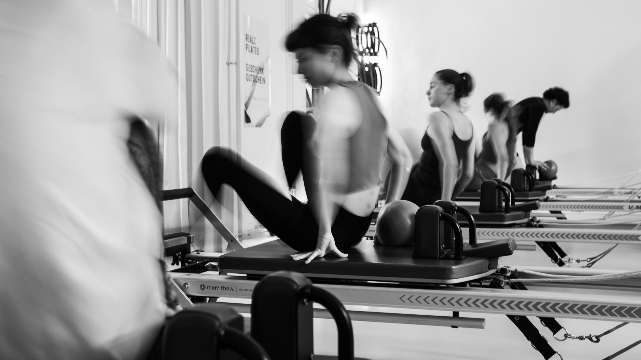 Eine Gruppe junger Klienten bereiten sich für ihre Pilates Gruppen Stunde im Pilates Studio REALZ Pilates vor. Sie sitzen auf dem Pilates Reformer. Die Klasse ist von Frauen und Männern gemischt.