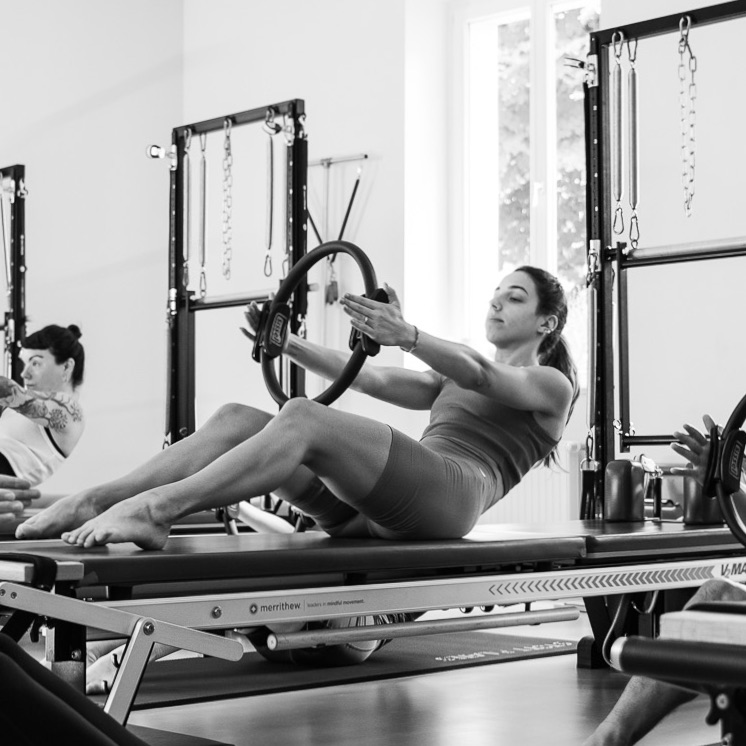 Eine junge Frau trainiert an der Pilates Tower Unit Wall. Die Arme sind mit dem Magic Circle lang ausgestreckt, ihr Schultern zentriert und entspannt. Eine Schulter-Kräftigung im Pilates. 