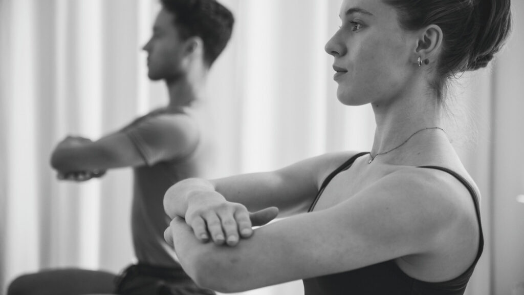 Eine junge Frau und ein junger Mann trainieren gemeinsam in der Pilates Chair Duo Klasse. Sie sitzen auf dem Pilates Chair, der Körper aufgerichtet, die Arme vor ihrer Brust über Kreuz, der Blick nach vorn. Sie sind sehr konzentriert in der Pilates Übung.