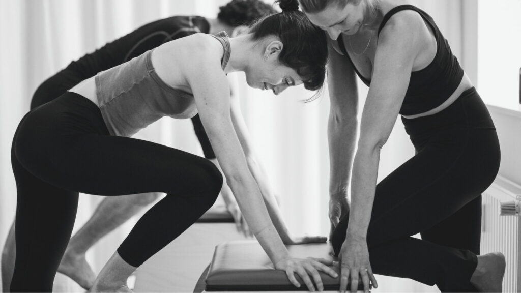 Das Pilates Wunda Chair Training im REAL Pilates Studio in Berlin. Ein Mann und eine Frau genießen das exklusive Duo Training. Sie stehen auf dem Chair Pedal, der Körper ist konzentriert und kraftvoll nach vorn gerichtet. Gemeinsam trainieren sie bedacht in Anleitung mit einer Pilates Lehrerin.