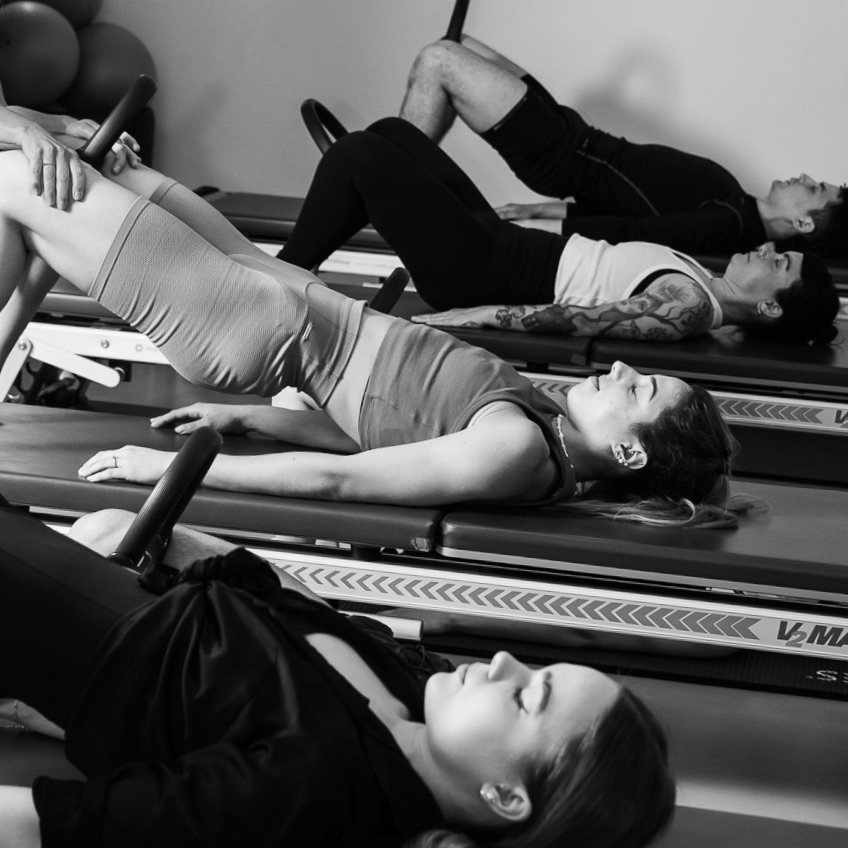 Pilates Gruppen Training an der Tower Unit Wall. Sie führen die Pilates Übung Bridging mit dem Pilates Magic Circle zwischen den Knien aus, um gezielt die Kraft aufzubauen.